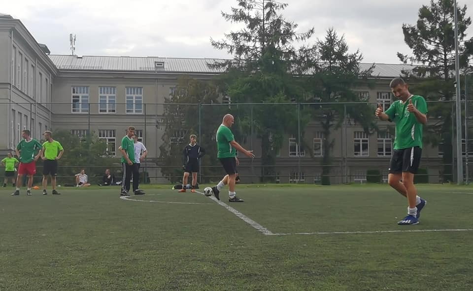 Na zdjęciu widoczny zespół FC 8 Wilki, rozgrzewający się na płycie boiska orlika. Po prawej stronie zdjęcia zawodnik zwrócony twarzą w stronę obiektywu, unosi kciuki w geście "ok". 