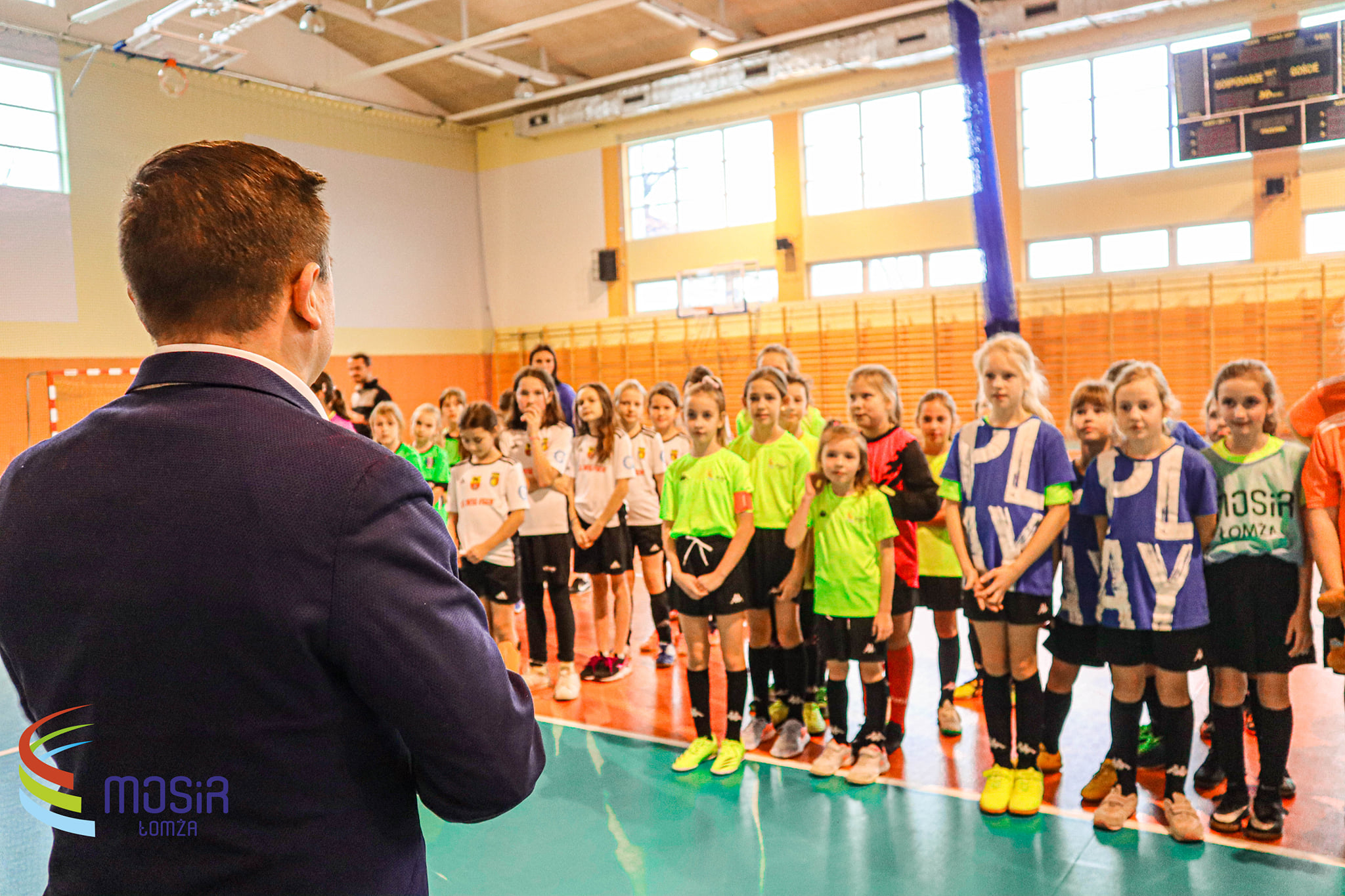 Ustawiony do obiektywu plecami w pierwszym planie widoczny Dyrektor MOSiR Łomża, na drugim planie około 60 dziewcząt w strojach sportowych. Zdjęcie wykonane na hali sportowej.