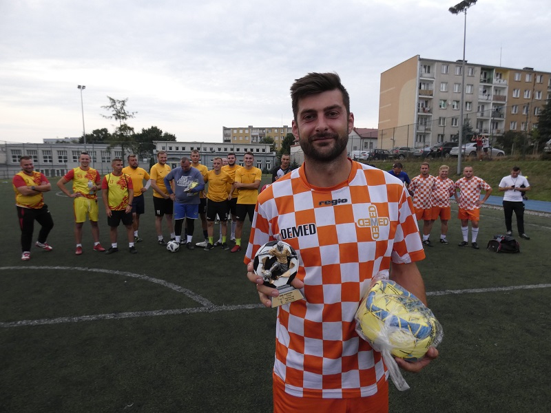 Na pierwszym planie stoi zawodnik w pomarańczowo-białej koszulce, trzymający w ręku piłkę i statuetkę za króla strzelców. W tle boisko i zawodnicy pozostałych zespołów.