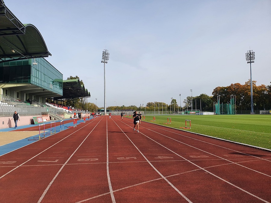 zdjęcie przedstawiające kilka osób biegnących po bieżni stadionu, w tle murawa, budynek stadionu, lampy