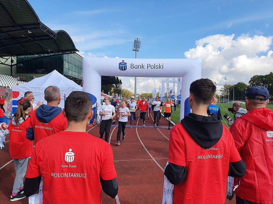 zdjęcie przedstawiające grupę osób biegnącą po bieżni stadionu i wolontariuszy stojących z medalami do wreczenia na mecie