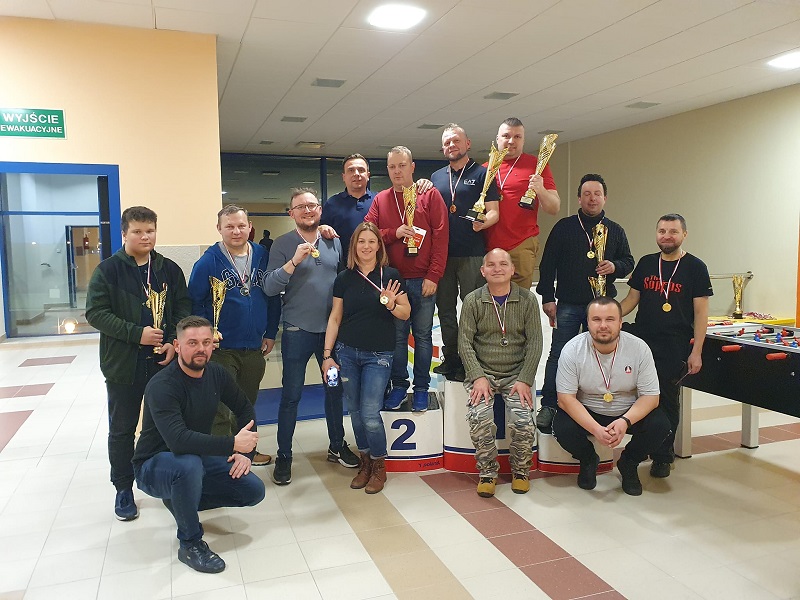 zdjęcie przedstawiające uczestników mistrzostw Łomży w piłkarzyki, z medalami i pucharami na podium, w tle stół do gry, drzwi