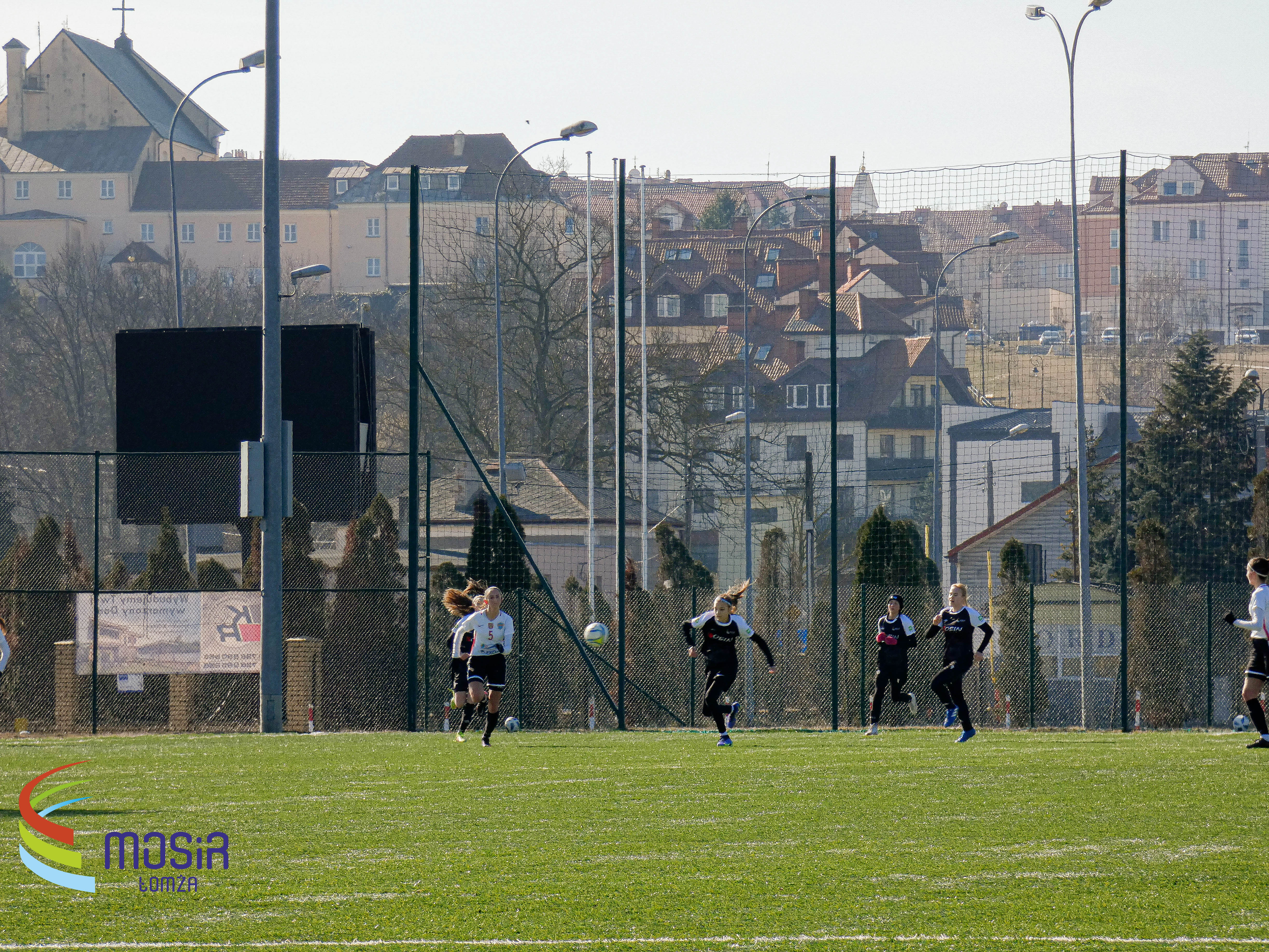 Boisko do piłki nożnej, na centralnej części fotografii dwie zawodniczki, jedna w czarnym, druga w białym stroju, w tle zabudowania miasta Łomża.