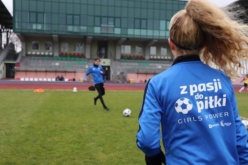 zdjęcie przedstawiające dwie zawodniczki żeńskiej druzyny piłki nożnej na murawie stadionu grające w piłkę, w tle budynek stadionu