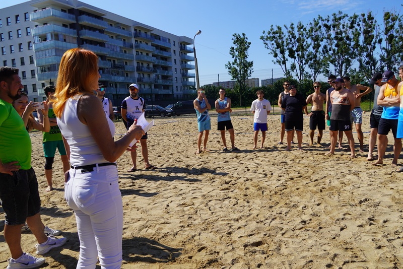 Zdjęcie przedstawia zawodników Turnieju Siatkówki Plażowej. Wszyscy są na powietrzu i stoją na piachu. Są ubrani w stroje sportowe.