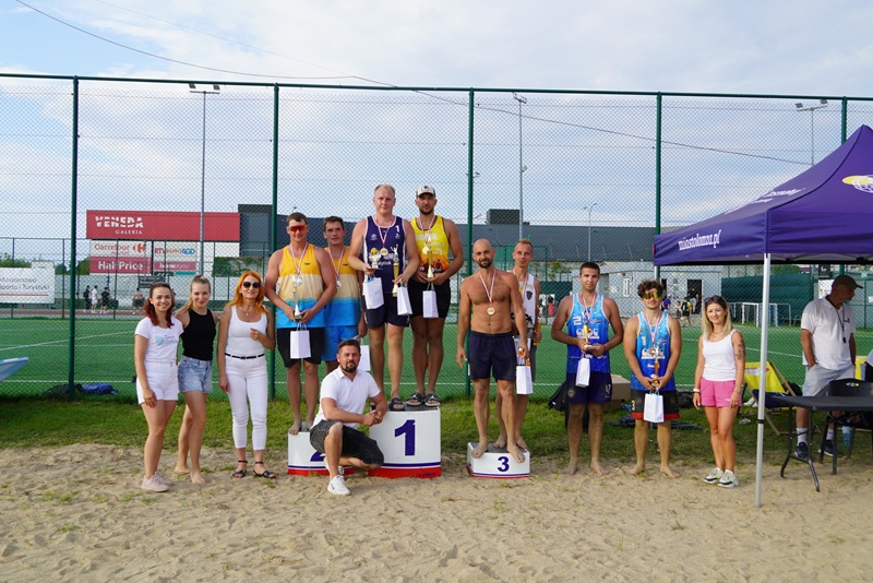 Zdjęcie przedstawia zwycięzców Turnieju w Siatkówkę Plażową. Wszyscy mają puchary i stoją na lub obok podium.